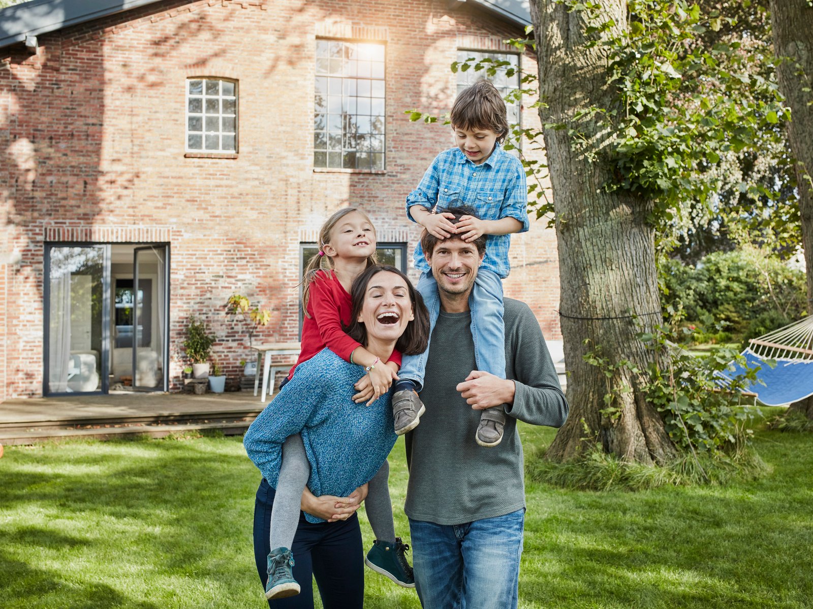 Familie mit vier Personen vor Backsteinhaus und Bäumen an sonnigem Tag.