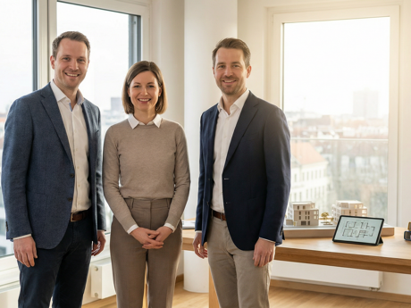 Drei lächelnde Personen im Büro mit Stadtblick und Modellen.