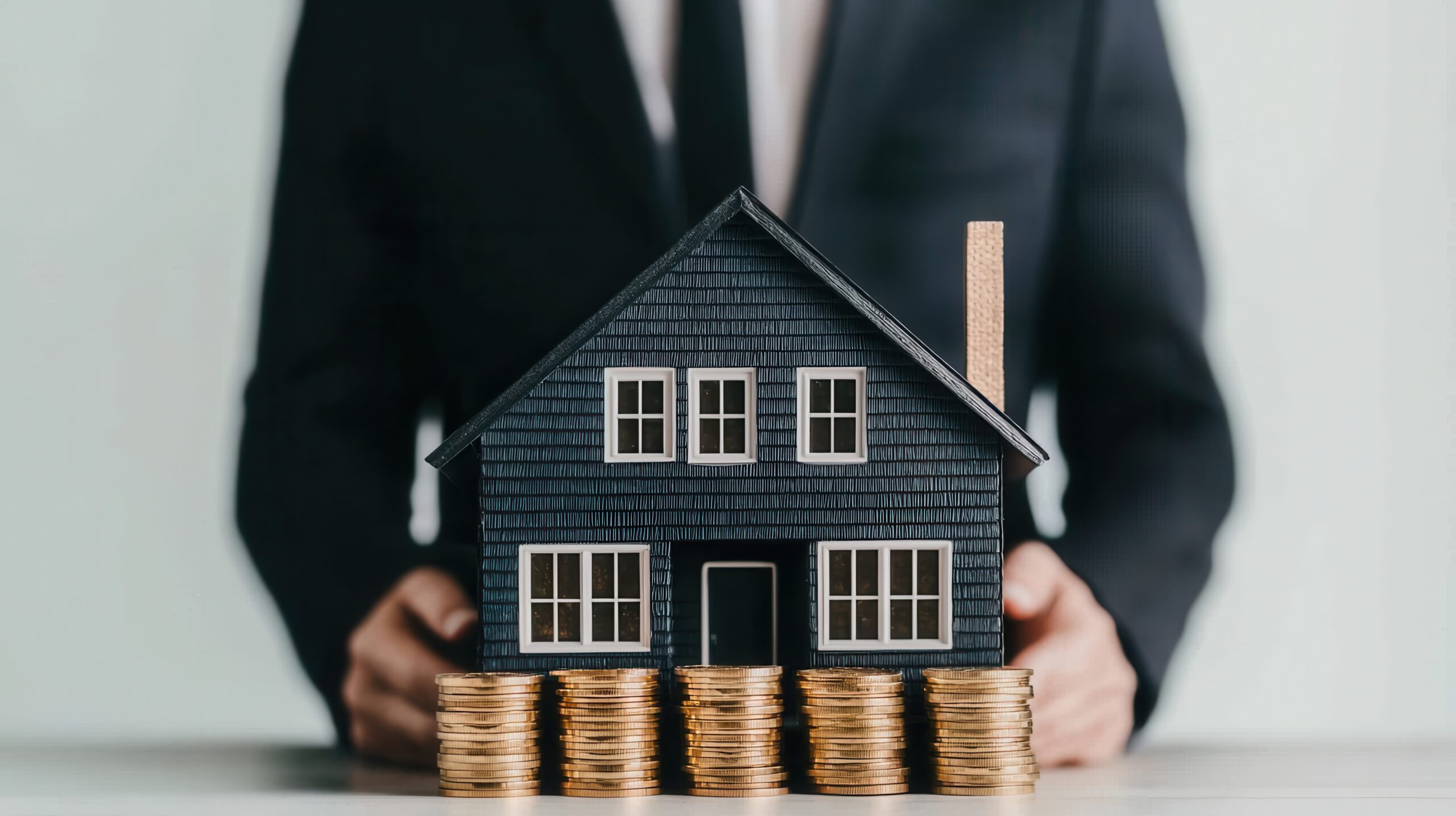 Person im Anzug mit Modellhaus und Münzstapeln davor.