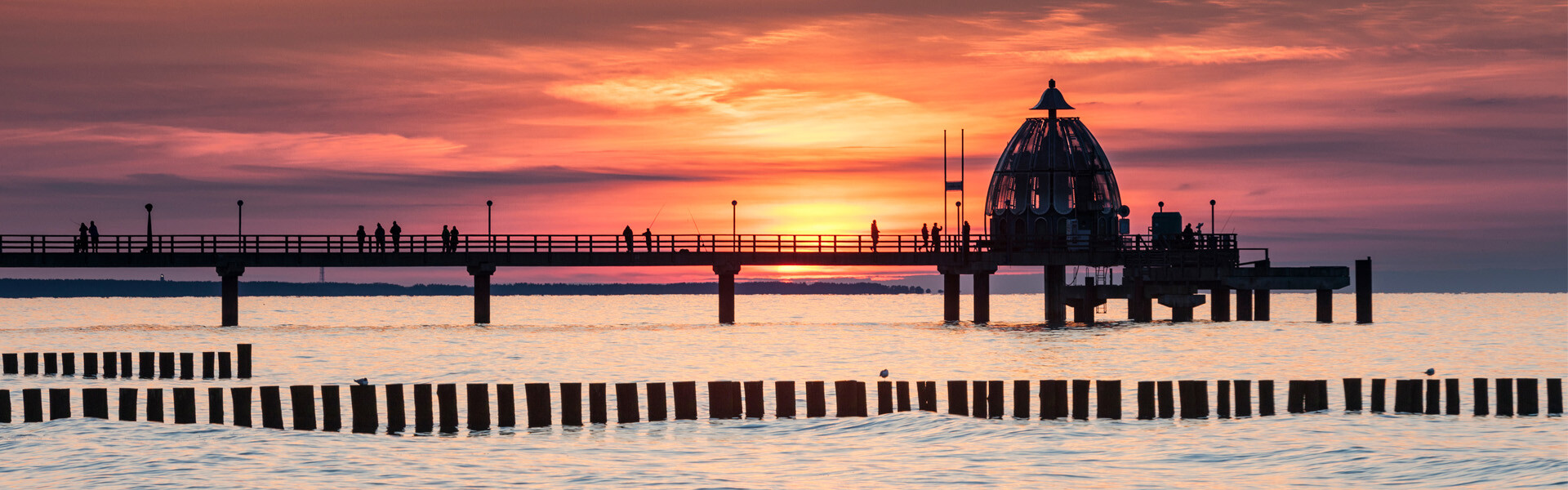 Pier bei Sonnenuntergang, Menschen als Silhouette vor orange-violettem Himmel.