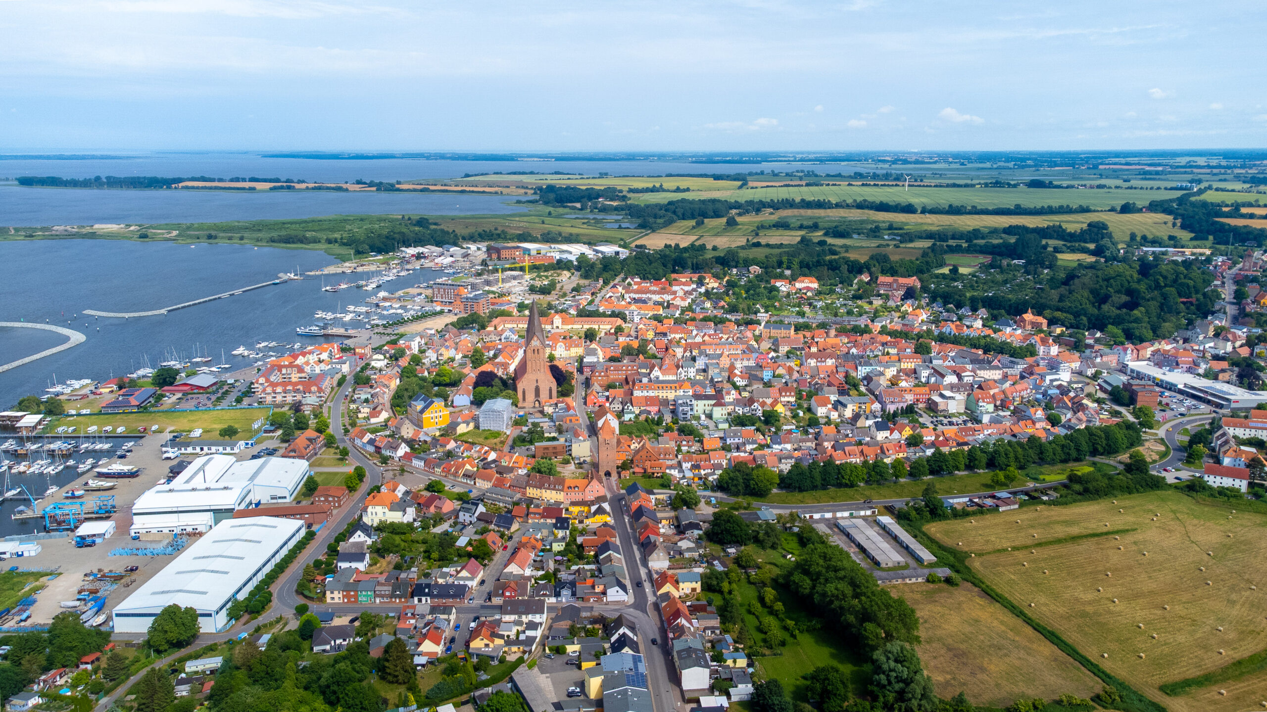 Küstenstadt mit bunten Häusern, Kirche, Hafen und grünen Feldern.