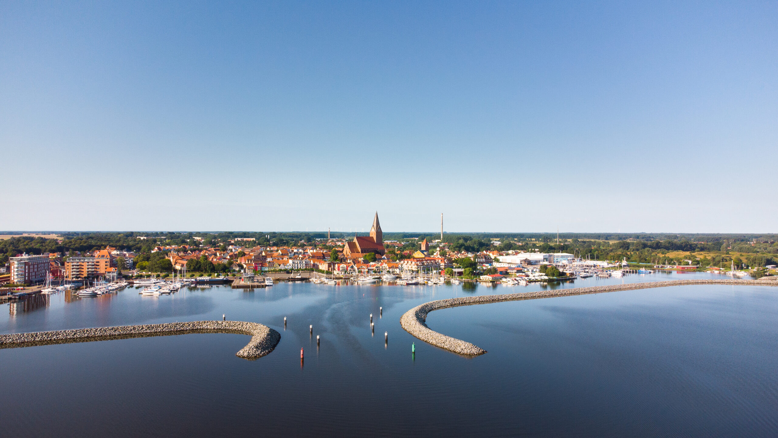 Küstenstadt mit Yachthafen, Wellenbrechern und hoher Kirchturmspitze.