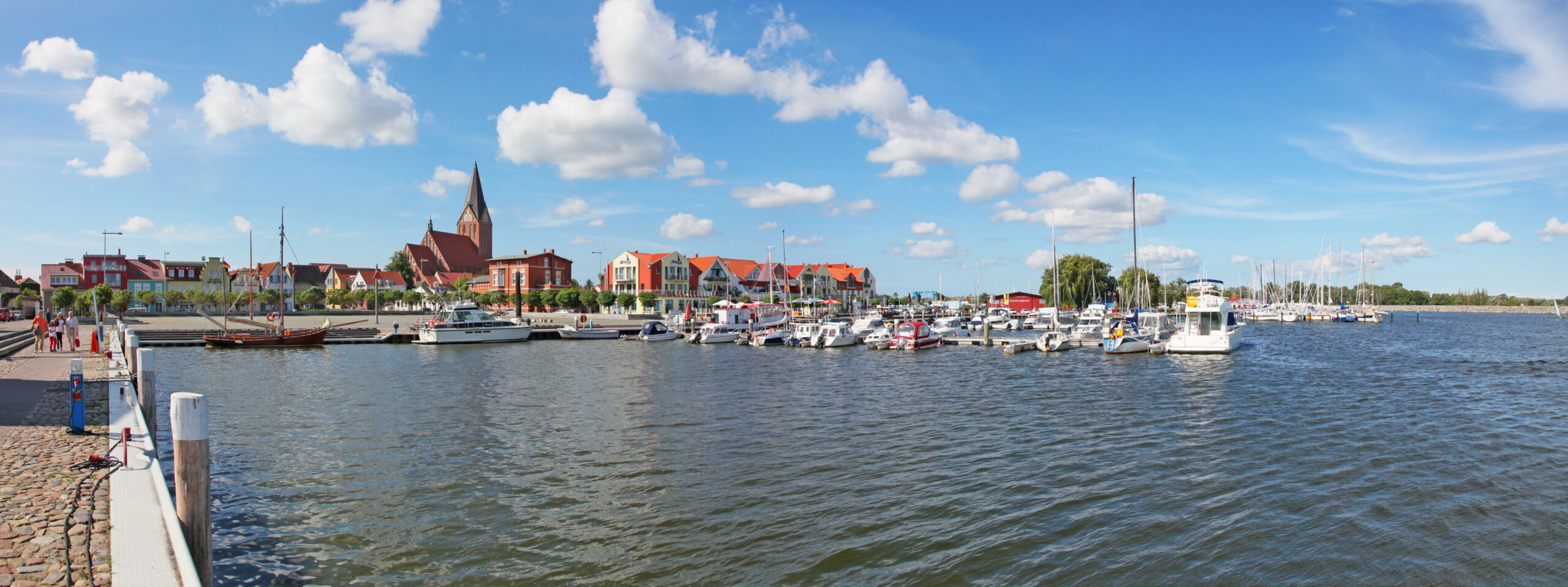Boote im Hafen, rote Dächer und Kirchturm bei blauem Himmel.