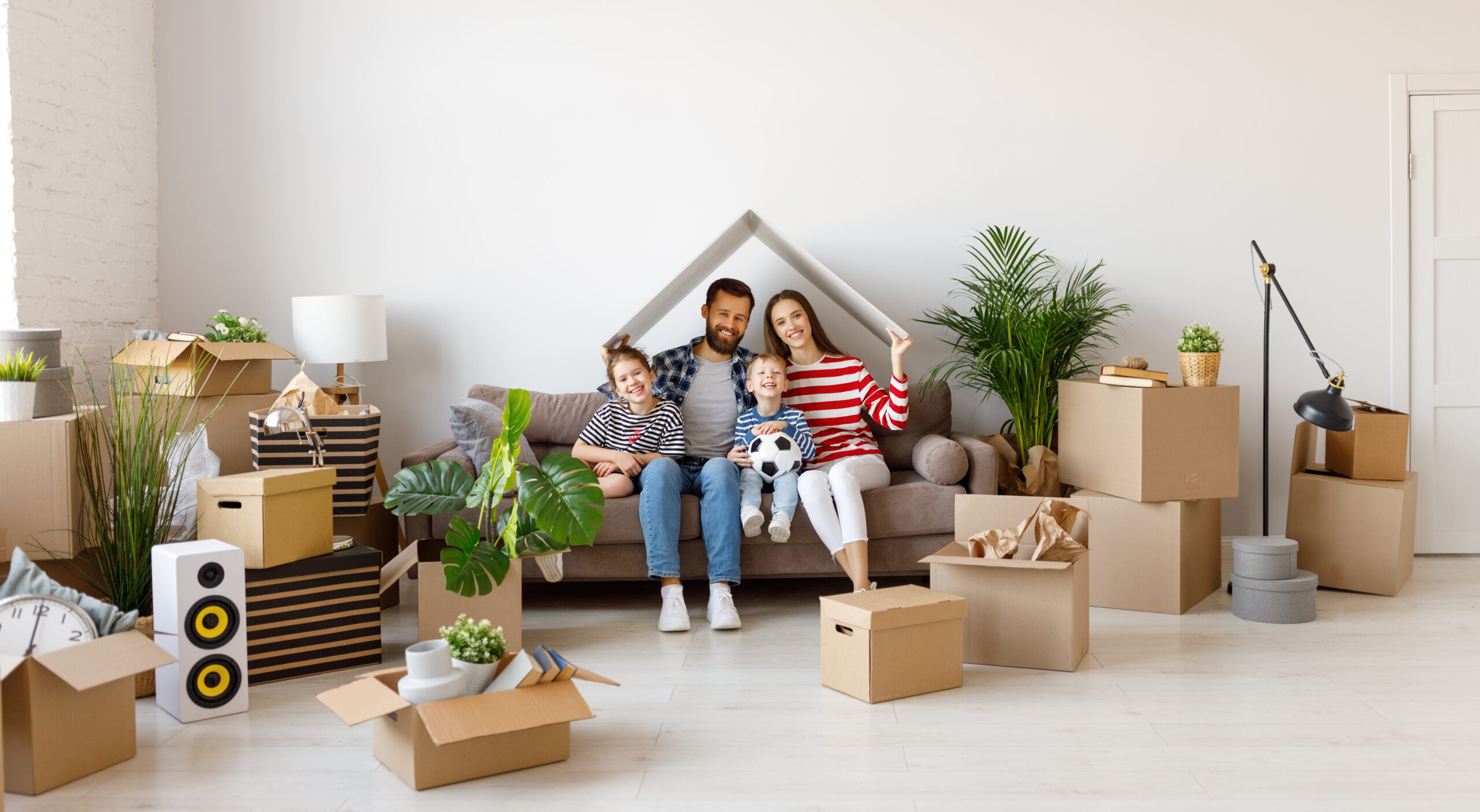 Lächelnde Familie mit Kindern auf der Couch inmitten von Umzugskartons und Pflanzen.